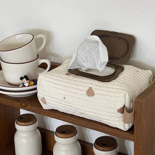 Quilted cream cotton tissue and wipe dispenser with brown accents, open with a wipe pulled out, on a wooden shelf.