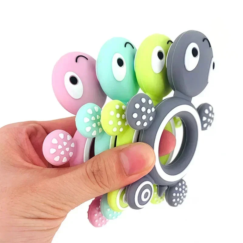 Hand holding four colorful silicone turtle teethers in pink, mint, lime, and gray with textured feet.