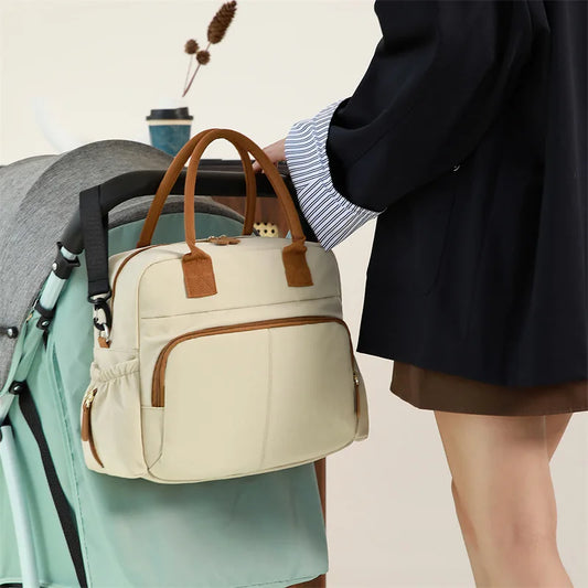 Stylish beige diaper bag with brown handles, attached to a stroller, held by a person.