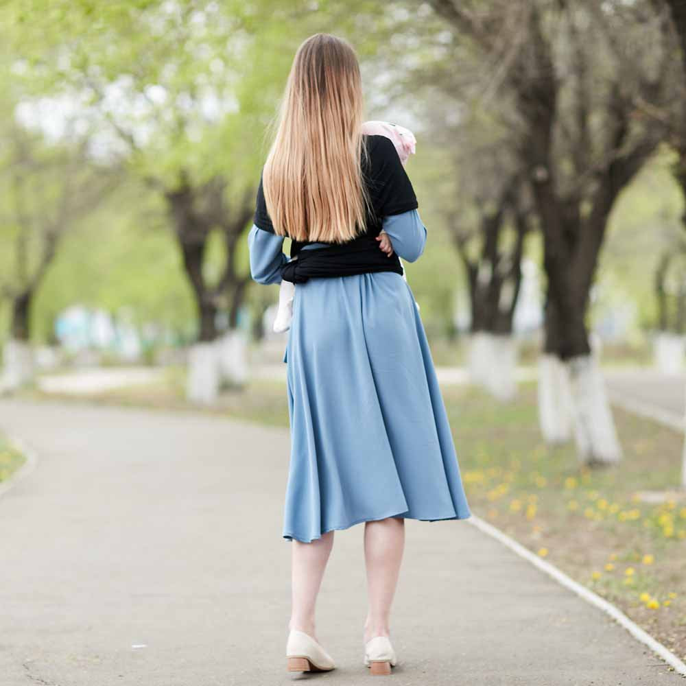 Woman from behind wearing a black fabric baby carrier with a light blue dress, walking on a path.