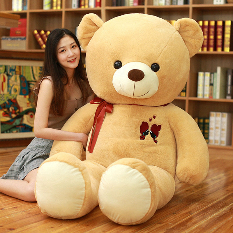 Large plush light brown teddy bear with a red ribbon and embroidered hearts, next to a smiling woman.