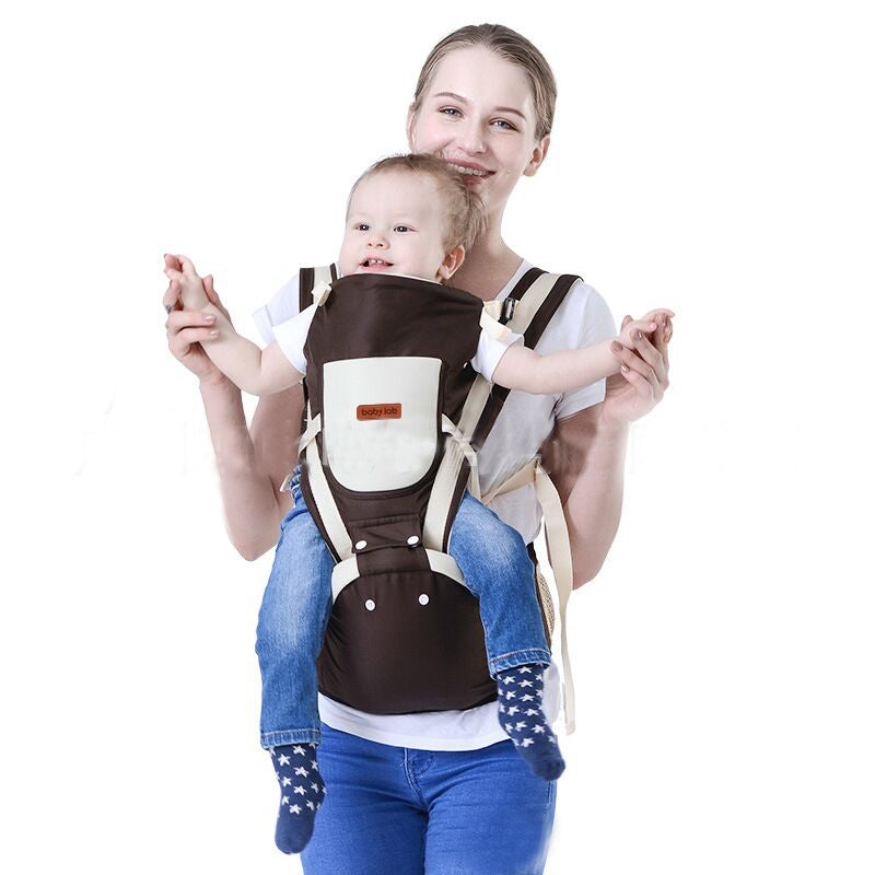 Smiling mother carrying baby in a brown and beige ergonomic baby carrier with hip seat.