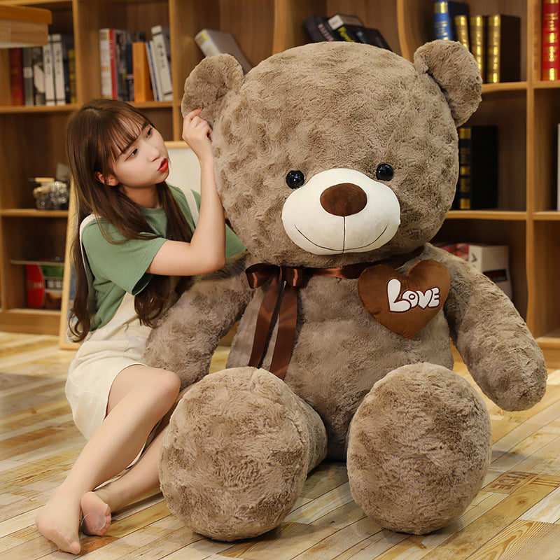 Giant soft brown teddy bear with a "Love" heart and ribbon, next to a young woman.