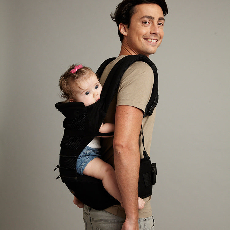 Smiling man carrying baby in black ergonomic back carrier with waist seat.