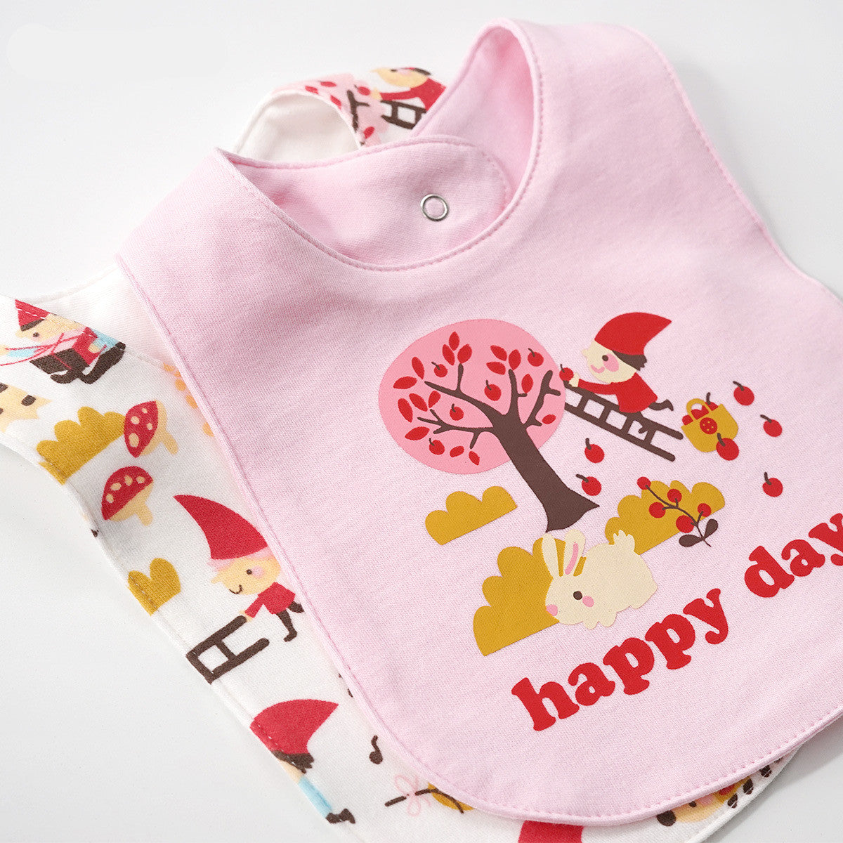 Pink and white cotton baby bibs with "happy day" text and gnome, tree, and bunny illustrations.