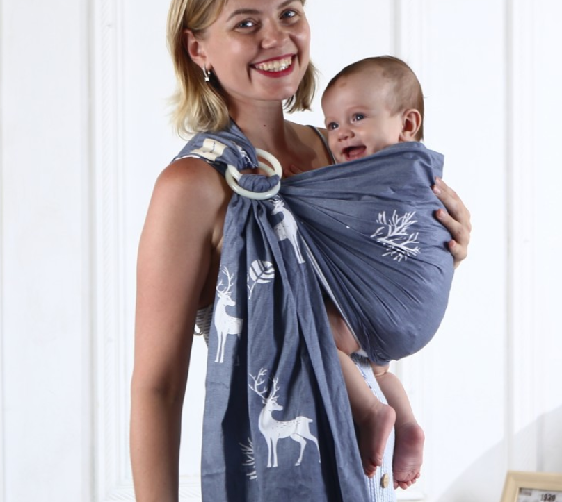 Smiling mother carrying happy baby in a soft, breathable blue cotton sling with white deer and tree patterns.