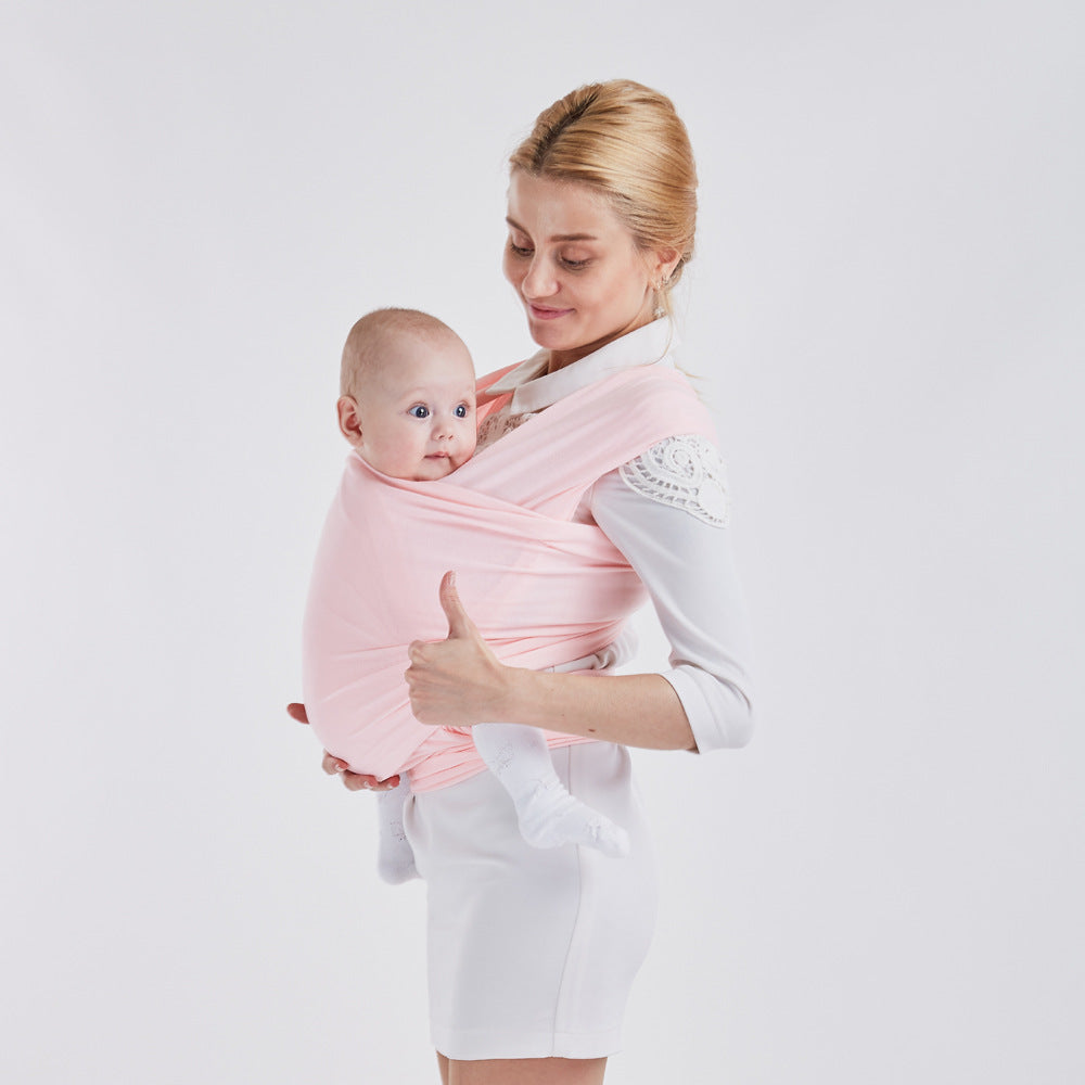 Smiling mother holding baby in a soft pink cotton baby wrap carrier, giving a thumbs-up.