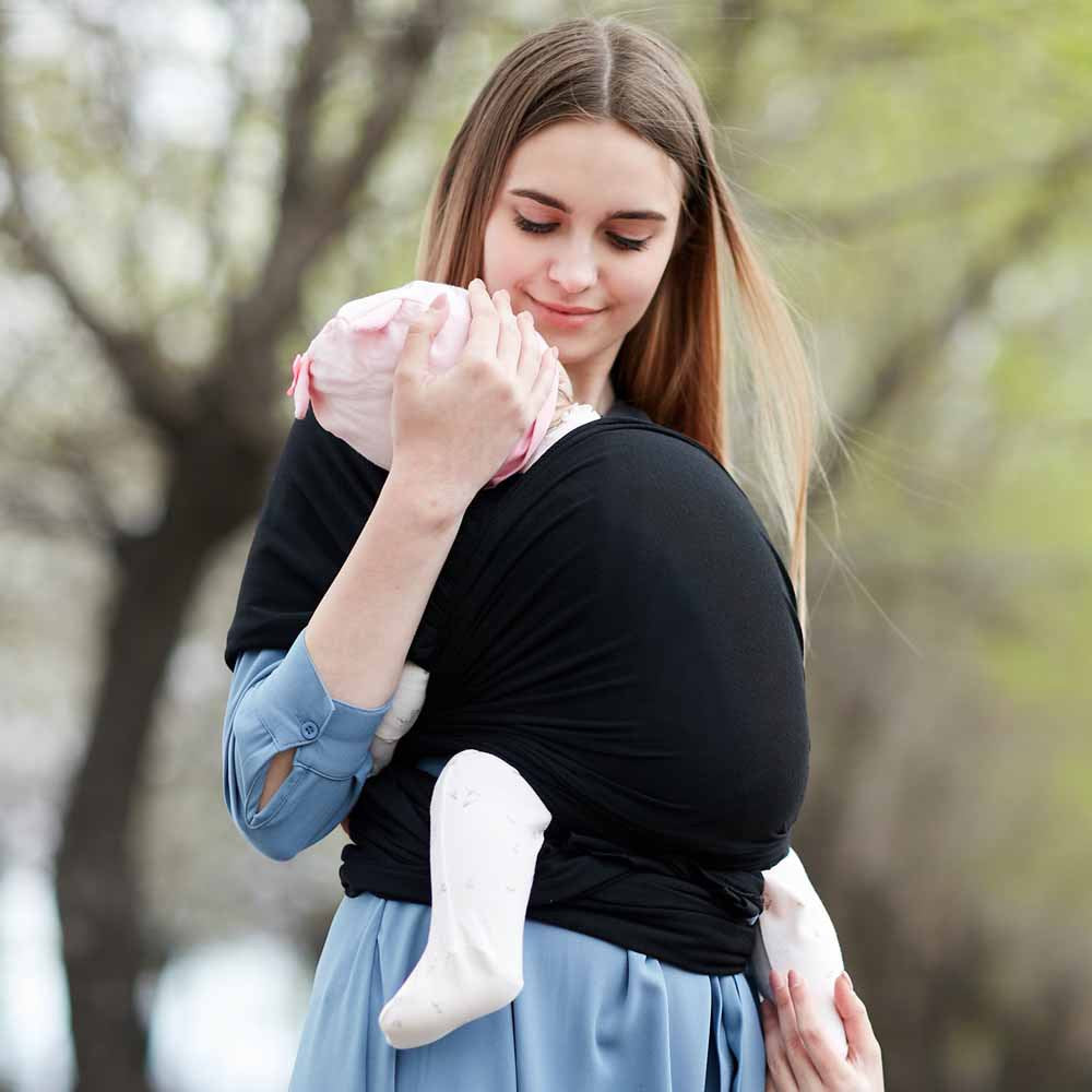 Young woman holding a baby in a black soft, breathable cotton baby wrap carrier outdoors.