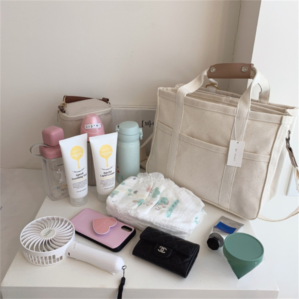 Beige canvas diaper bag with brown leather handles, surrounded by baby essentials and personal items.