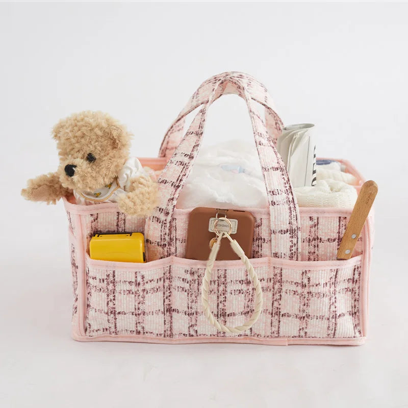 Pink plaid fabric diaper caddy with handles, filled with baby essentials like a teddy bear, diapers, and accessories.