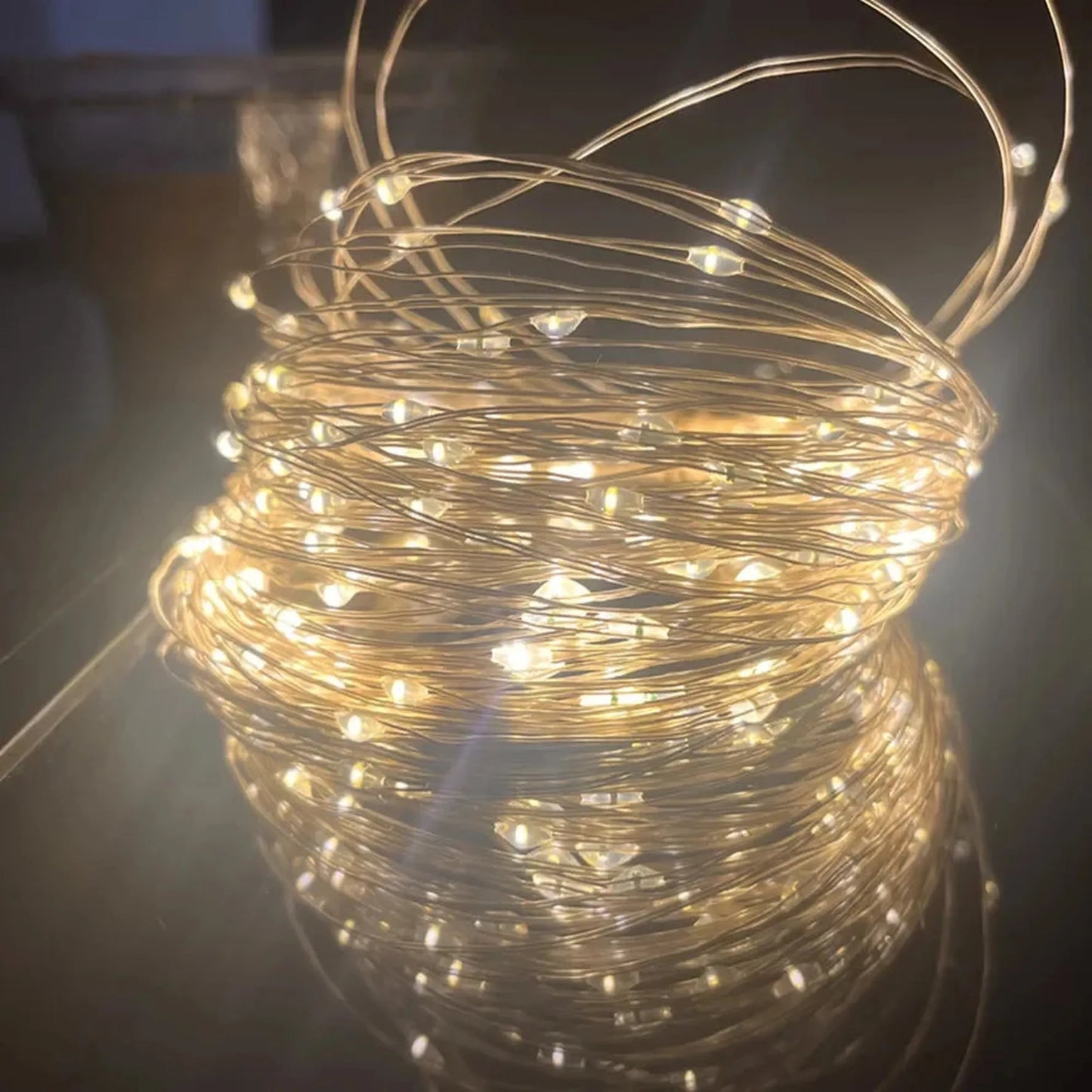 Warm white LED string lights on a flexible copper wire, coiled and glowing on a reflective surface.