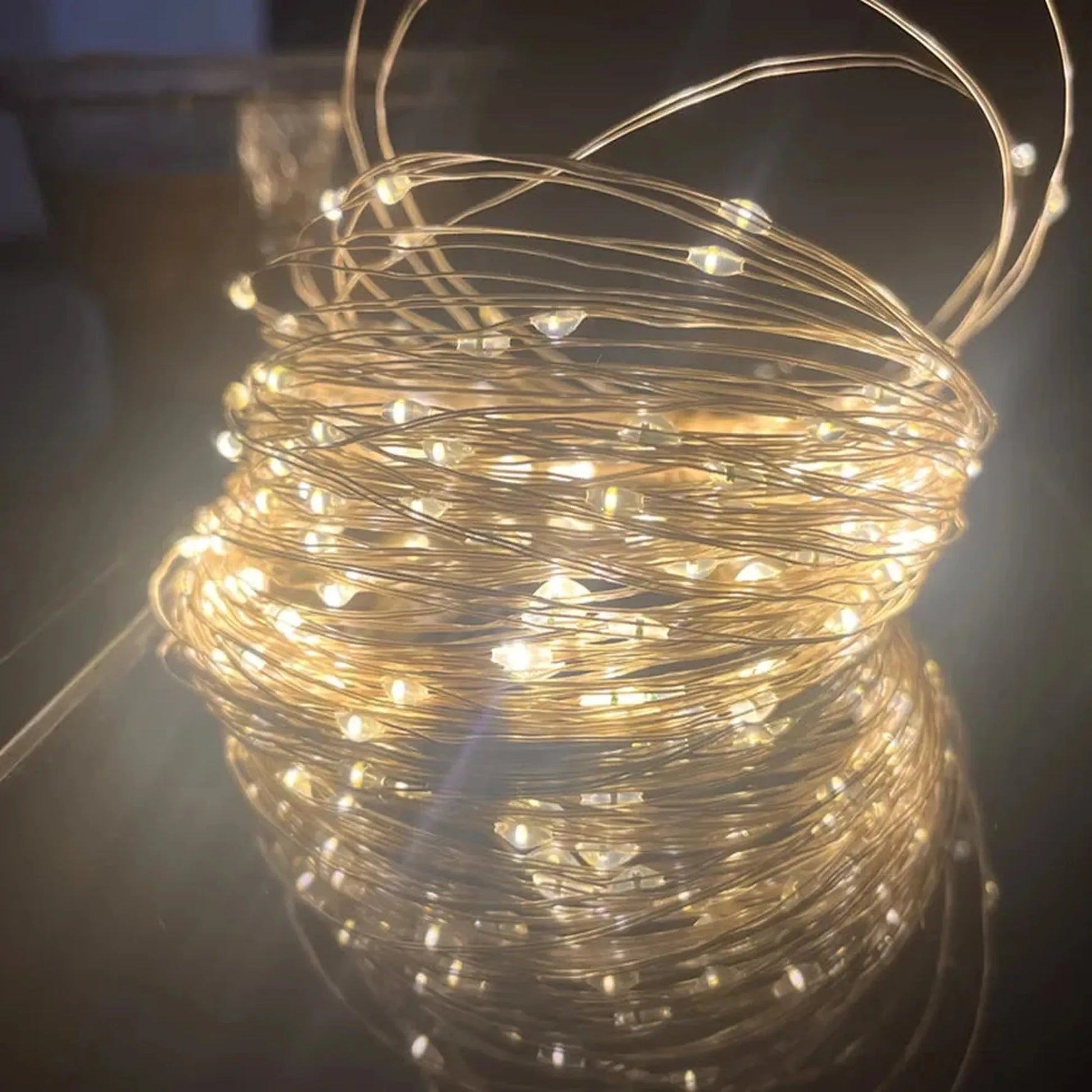 Warm white LED string lights on a flexible copper wire, coiled and glowing on a reflective surface.