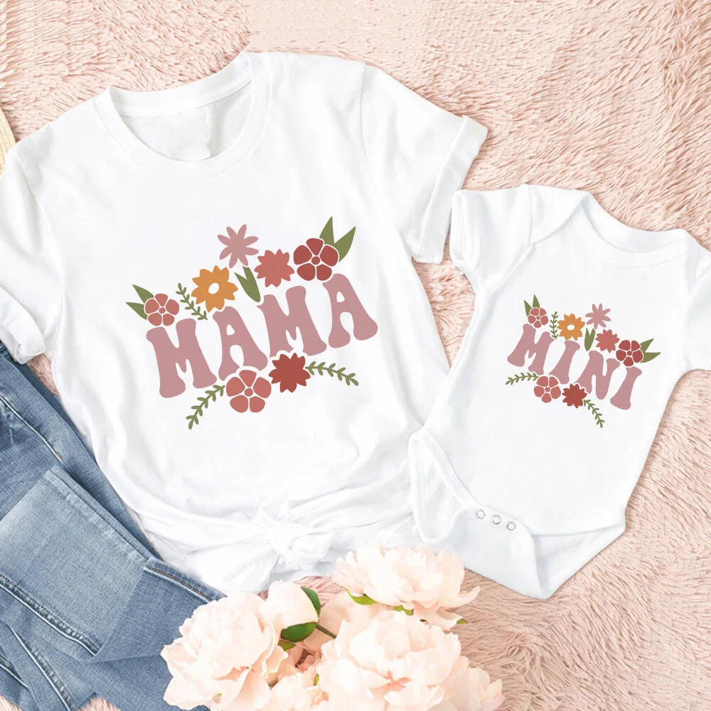 White "Mama" t-shirt and "Mini" baby bodysuit with matching floral designs, next to jeans and flowers.