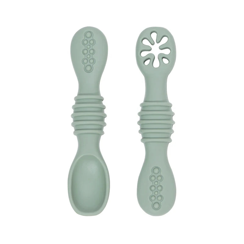 Two soft sage green silicone baby training spoons, one solid and one with a flower-shaped feeder.