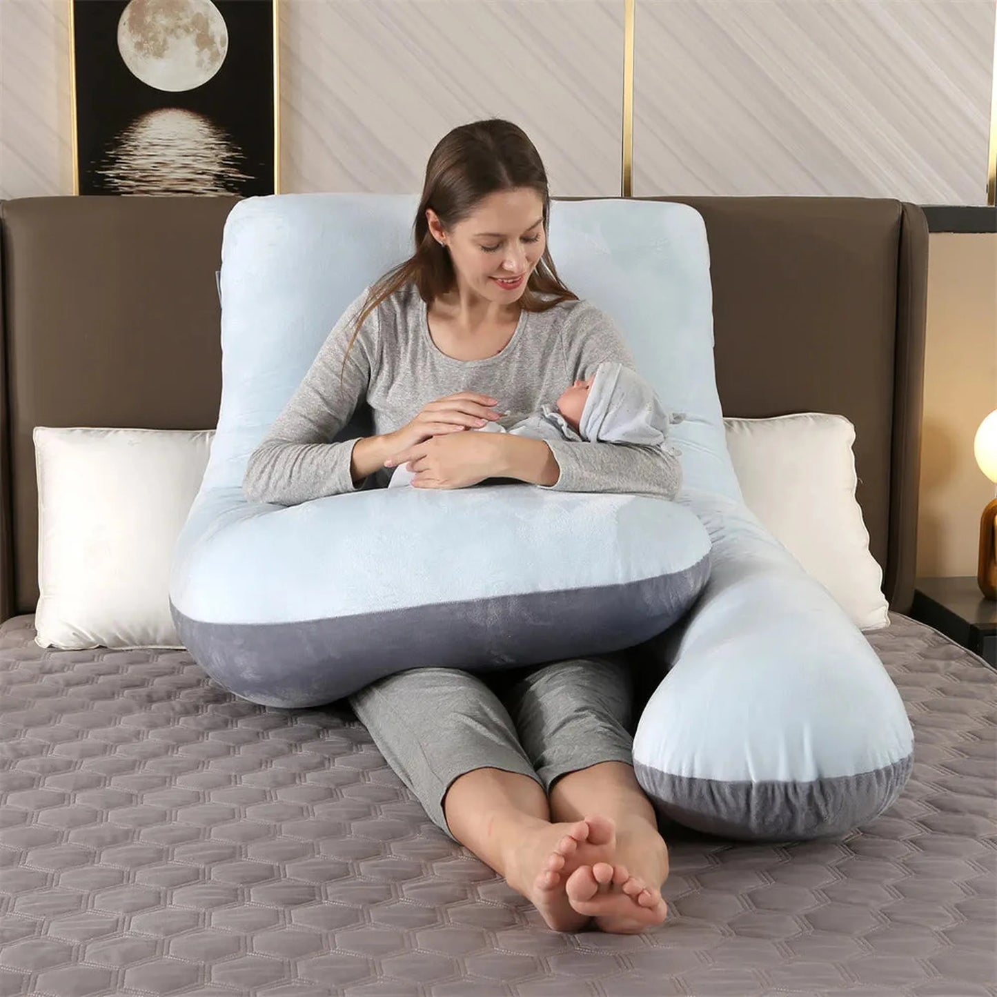 Woman holding a baby while sitting comfortably with a full-body gray and light blue orthopedic pregnancy pillow.