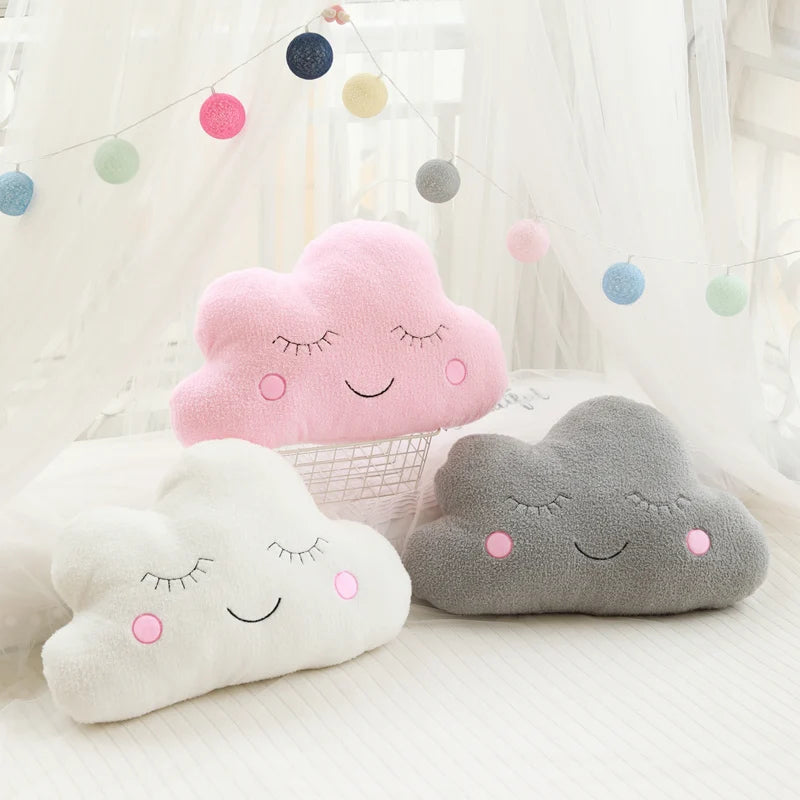 Soft plush cloud pillows in pink, white, and gray with embroidered sleepy faces for nursery decor.