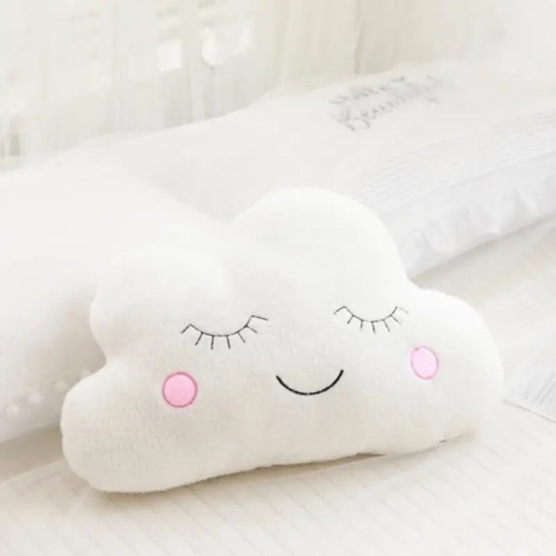 Soft white cloud-shaped plush pillow with embroidered sleepy eyes and pink rosy cheeks, ideal for nursery decor.
