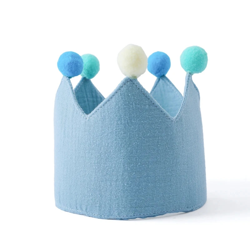 Light blue cotton baby birthday crown with colorful blue, teal, and cream pom-poms on a white background.