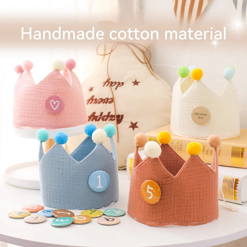 Four handmade cotton baby birthday crowns in pink, blue, cream, and rust, with colorful pom-poms and number badges.