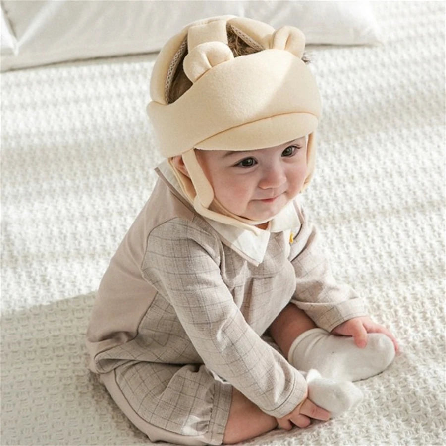 Cute baby wearing a beige, breathable anti-collision helmet with ear details, sitting on a white blanket.