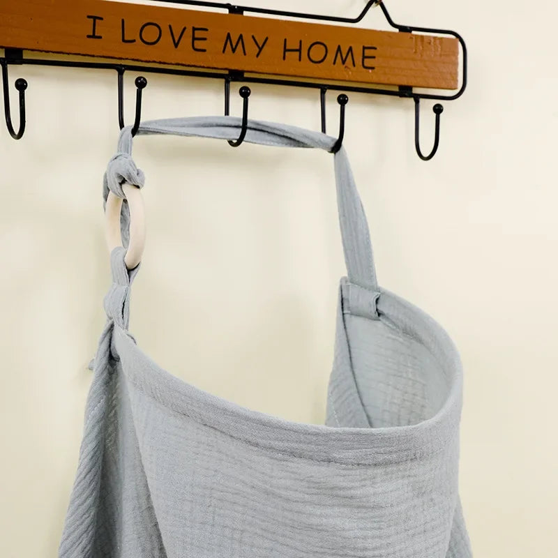 Light gray cotton nursing cover and stroller blanket with a wooden ring, hanging on a wall hook.