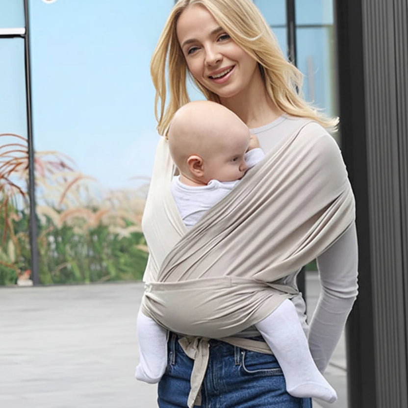 Smiling woman wearing a beige ergonomic soft cotton baby wrap carrier with a baby facing her.