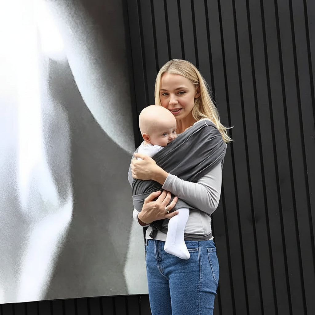 Blonde woman holding a baby in a dark gray ergonomic cotton baby wrap carrier.