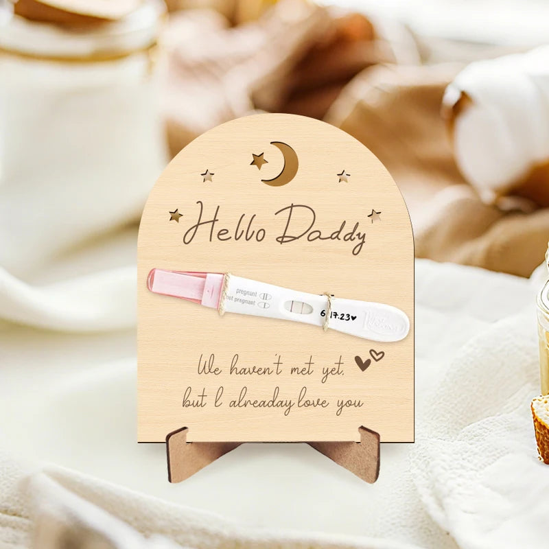 Arched wooden pregnancy announcement sign with "Hello Daddy" and "We haven't met yet but I already love you" text, holding a positive pregnancy test.