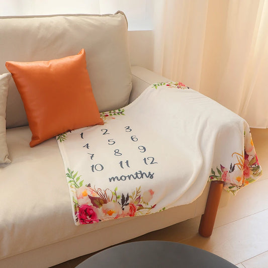 Soft white polyester baby milestone blanket with floral design and numbers 1-12, draped on a sofa.