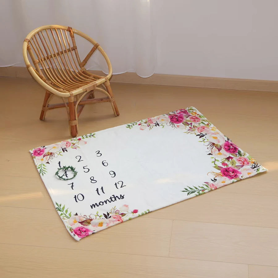 White polyester baby milestone blanket with pink floral border and numbers 1-12, next to a small rattan chair.