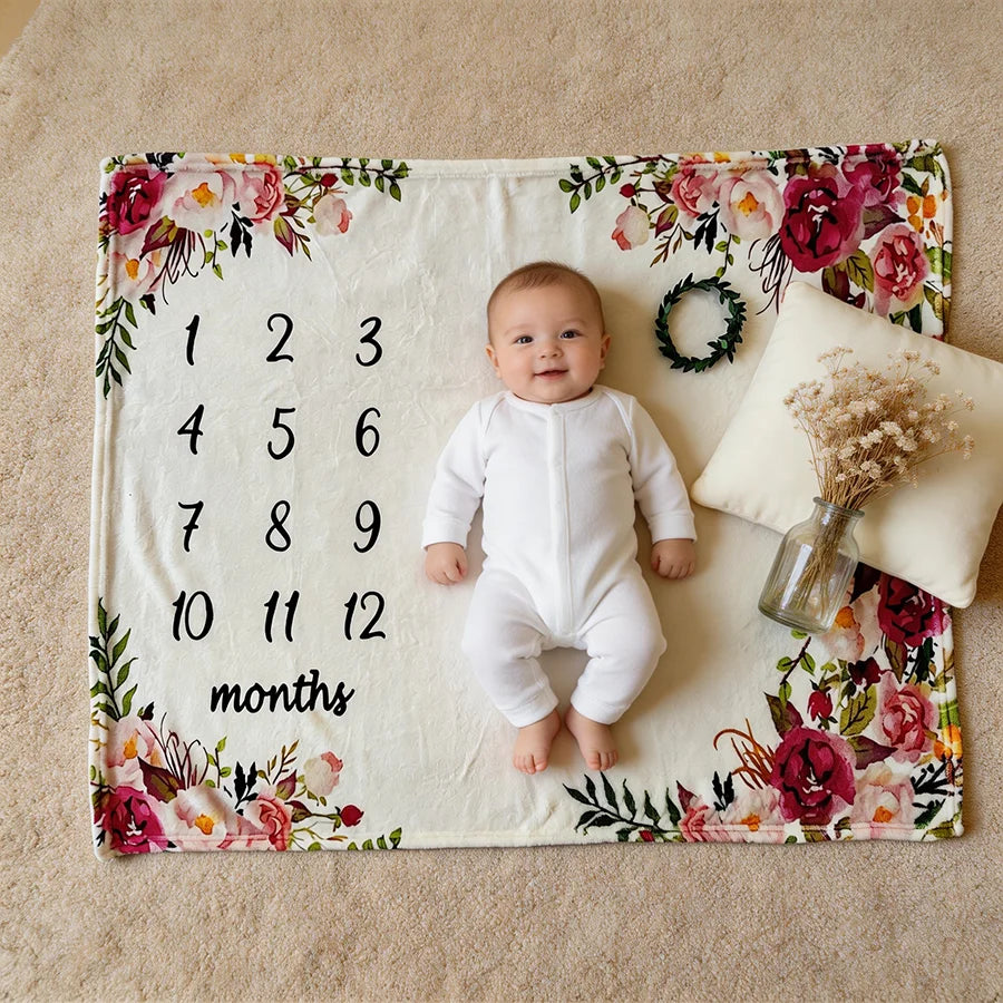 Baby on a white floral milestone blanket with numbers 1-12, a pillow, and dried flowers.