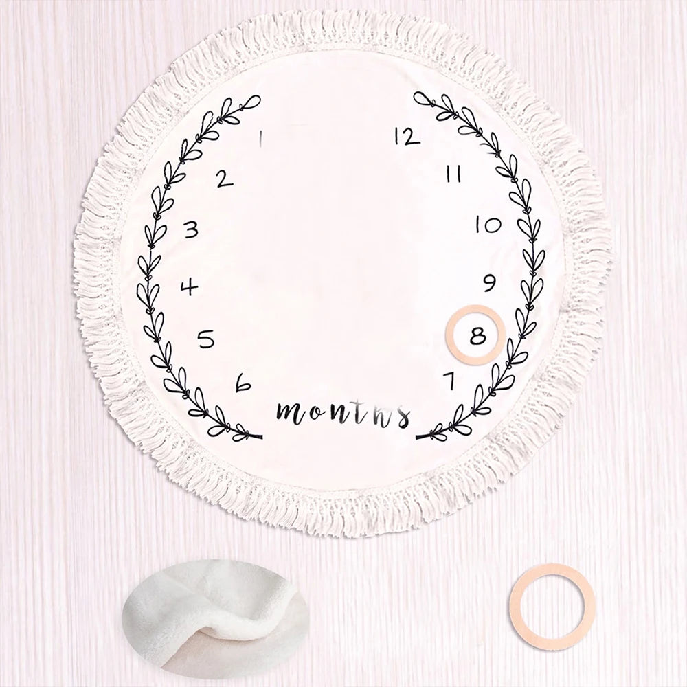 Round white flannel baby milestone blanket with fringe, leaf wreath, numbers 1-12, and two wooden markers.