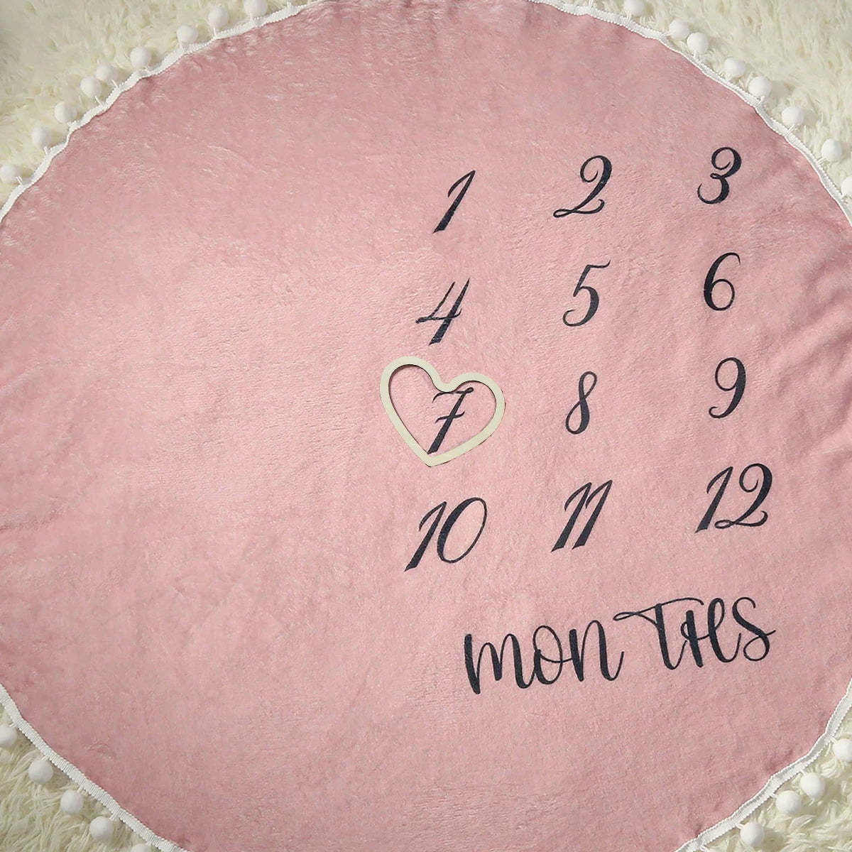 Soft pink flannel baby milestone blanket with monthly numbers, a heart marker, and pom-pom trim.