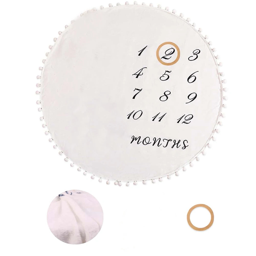White round flannel baby milestone blanket with black numbers 1-12, "MONTHS" text, and pom-pom trim.