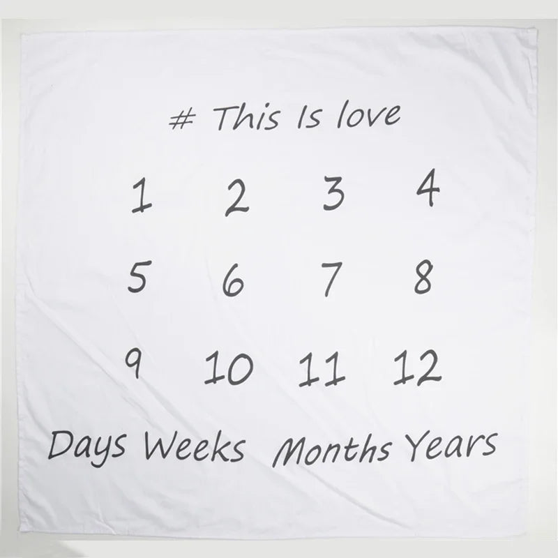 White polyester baby milestone blanket with "This Is love," numbers 1-12, and "Days Weeks Months Years" in black script.