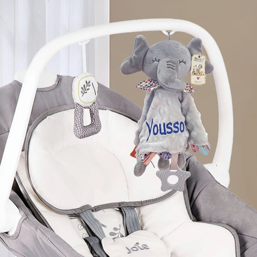 Soft gray elephant security blanket with personalized "Youssou" embroidery, colorful trim, and star teether, hanging on a baby bouncer.