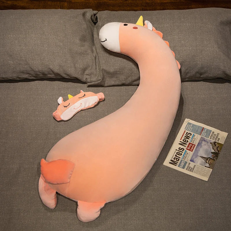Soft pink unicorn plush body pillow with yellow horn and a matching eye mask on a gray bed.