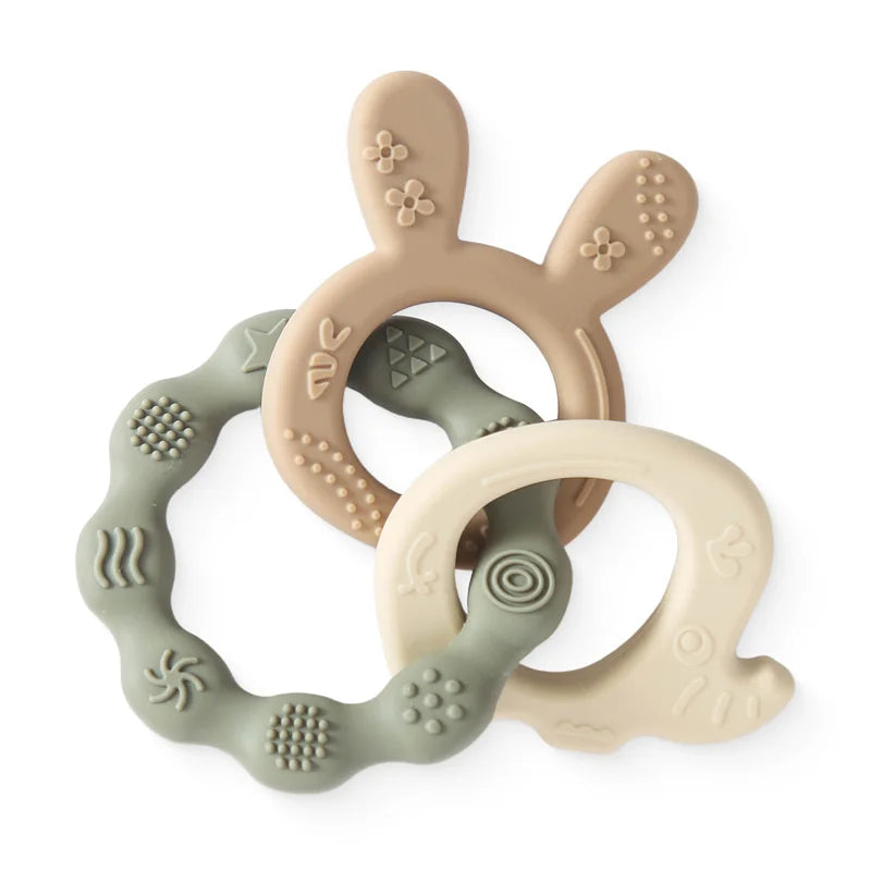 Three silicone baby teethers: a beige rabbit, a sage green wavy ring, and a cream wavy ring, all with textured surfaces.
