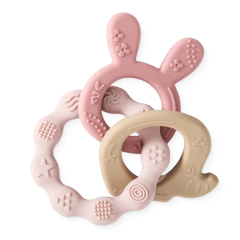 Three silicone baby teethers: a pink rabbit, a beige ring, and a light pink textured ring.