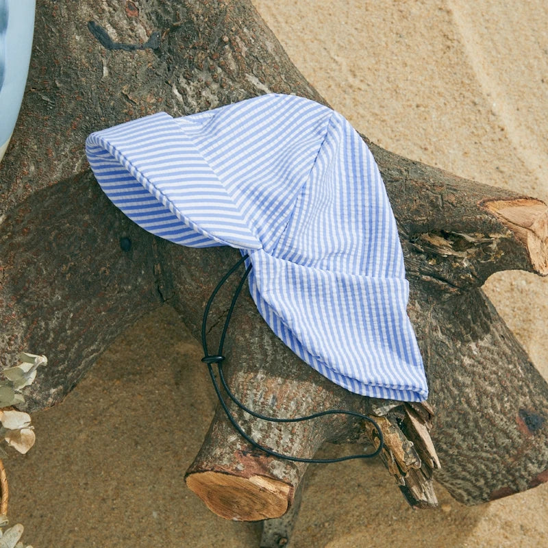 Blue and white striped baby sun hat with a chin strap, made from polyester and elastane, on a tree trunk.