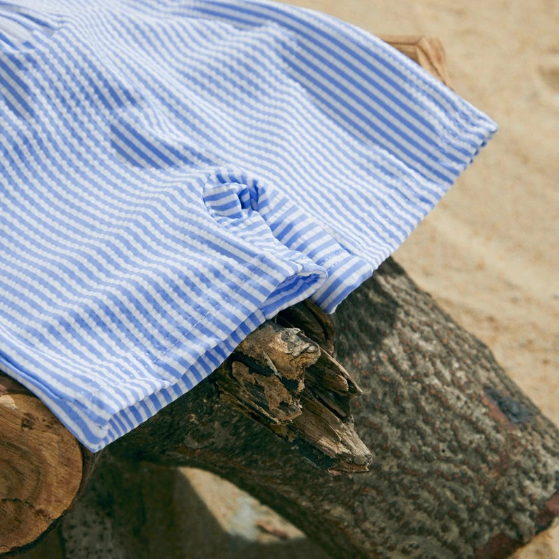 Blue and white striped short-sleeve baby swimsuit with a half-zip, lying on a log on a sandy beach.