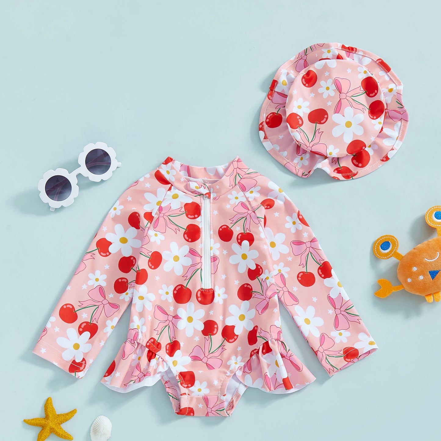 Pink long-sleeve rashguard swimsuit for babies with cherry and daisy print, matching hat, and front zipper.