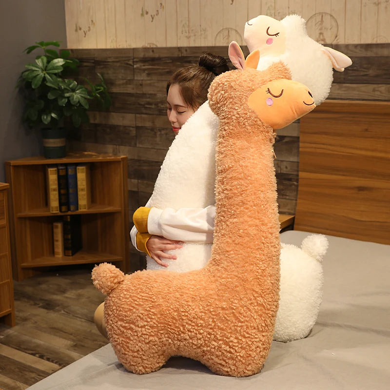 Soft brown alpaca plush toy with a long neck, hugged by a person on a bed.