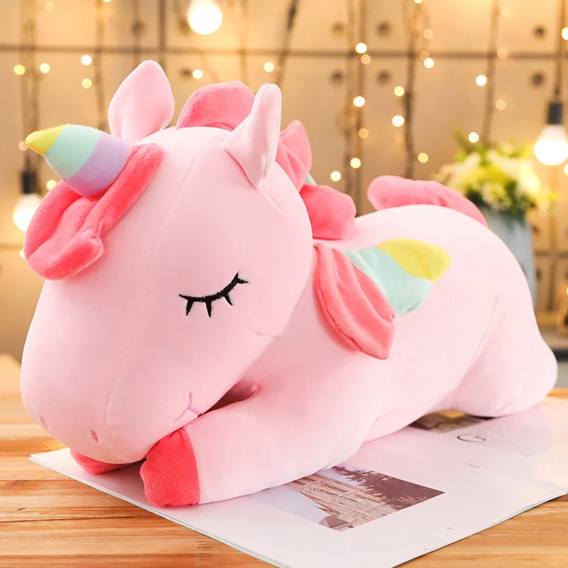 Soft pink plush unicorn toy with closed eyes, rainbow horn and wings, lying on a book.