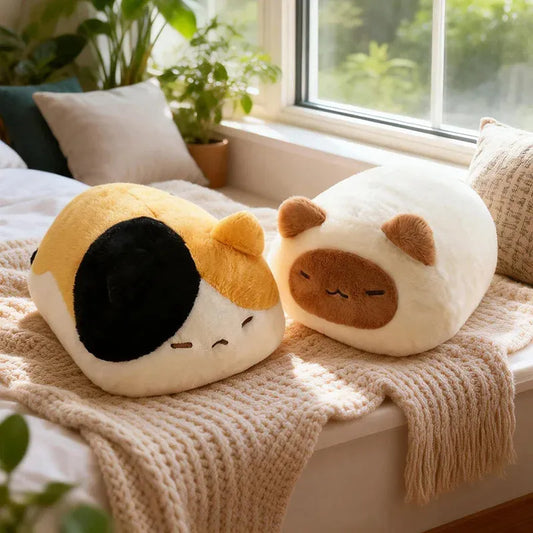 Two soft, plush cat pillows: one calico, one cream with brown ears, on a window seat.