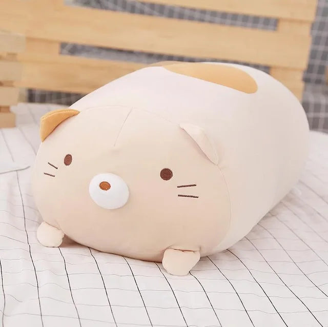 Soft, light brown plush cat pillow with small ears and embroidered facial features, resting on a checkered bed.