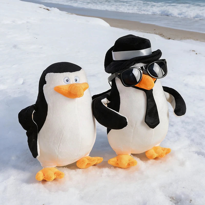 Two plush Madagascar penguins on snow; one wears a black hat, sunglasses, and tie.