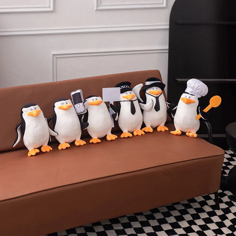 Six soft plush Madagascar penguins, 28cm, in various costumes on a brown sofa.
