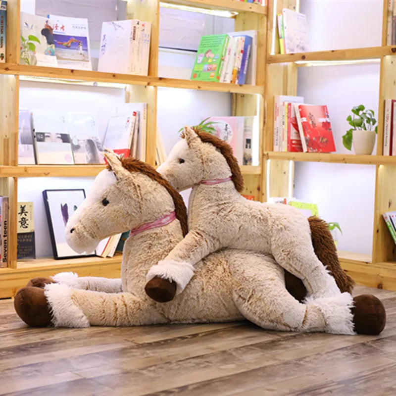 Two large, soft brown plush horse toys, one lying down with a smaller one on its back, against a bookshelf.