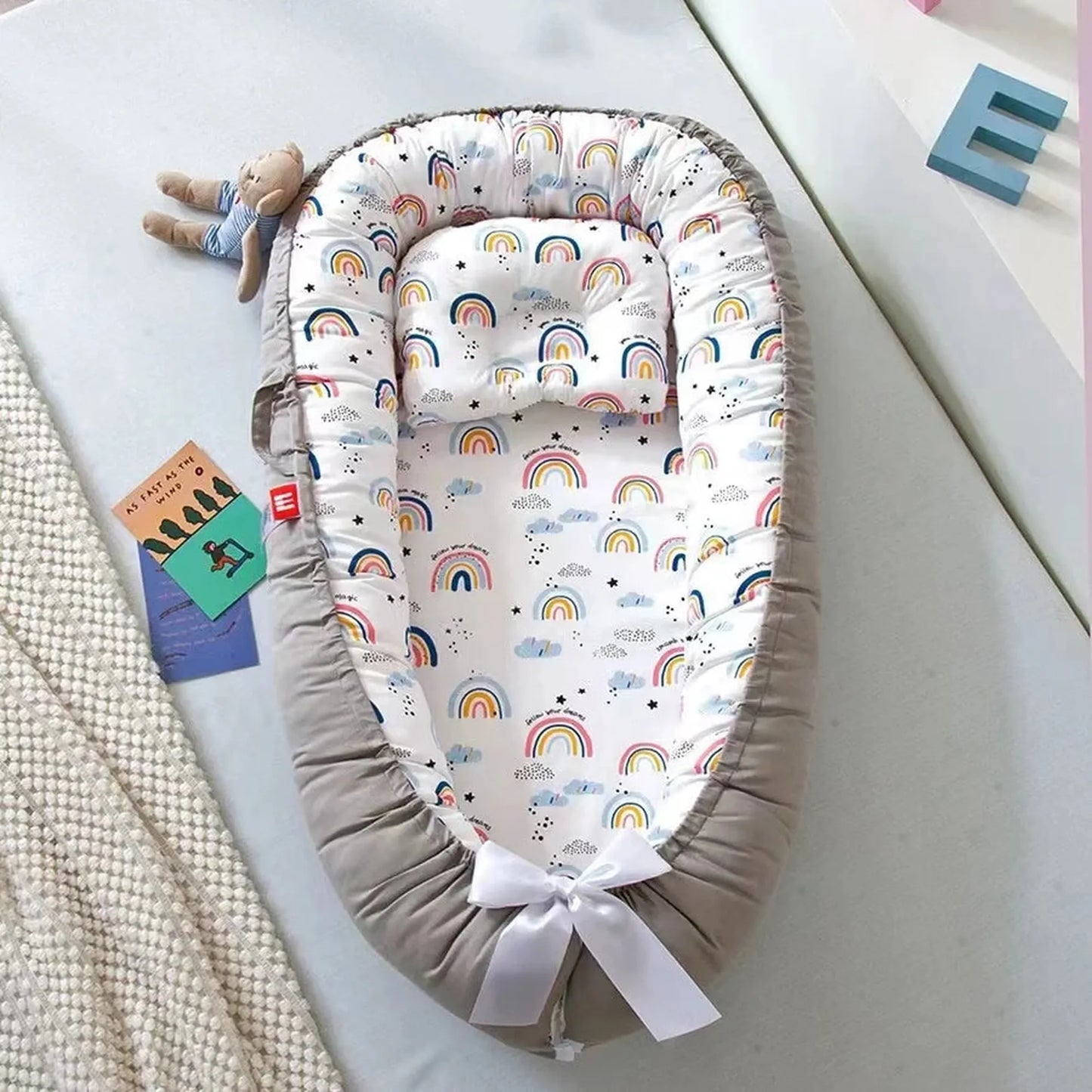 Breathable baby nest with rainbow print, adjustable gray cotton sides, and white bow detail.